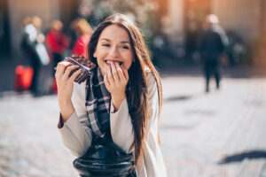 A young woman with long brown hair smiles as she enjoys chocolate outdoors, wearing a light coat and scarf, against a blurred city background—inviting you to eat more chocolate and savor the moment.