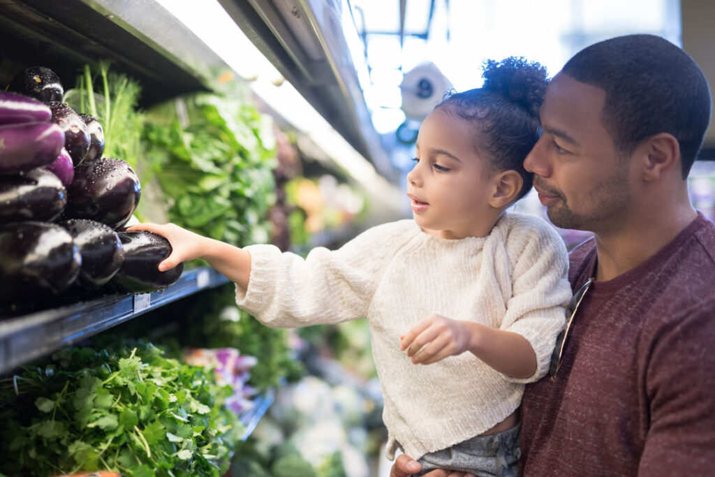 A pre-school age girl helps her dad pick out healthy food and veggies in the produce section at the grocery store.