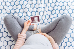 9 months pregnant young woman holding hand on her belly, she is sitting on bed and hold ultrasound image of baby