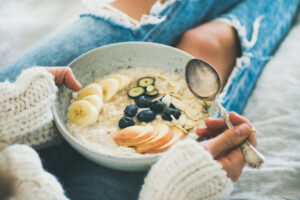 Healthy winter breakfast in bed. Woman in woolen sweater and shabby jeans eating vegan almond milk oatmeal porridge in bowl with berries, fruit and almonds. Clean eating, vegetarian food