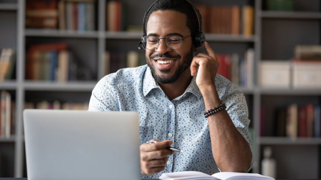 Smiling African American man in glasses and headset watch webinar on laptop making notes, happy biracial male student worker in headphones handwriting studying or working using computer