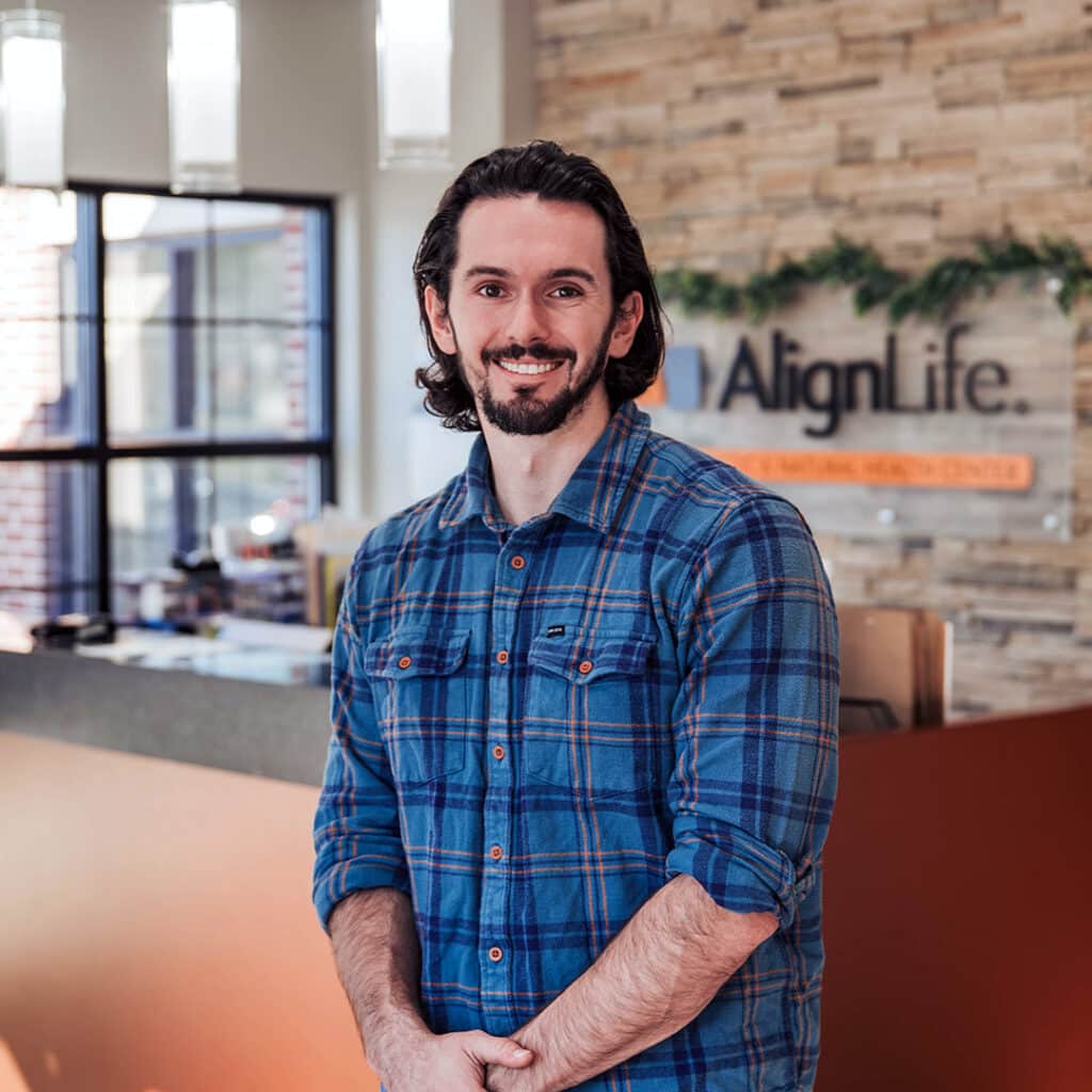 A man with long dark hair and a beard, wearing a blue plaid shirt, stands in front of the reception desk at AlignLife Natural Health Center, a trusted Fishers Chiropractor.
