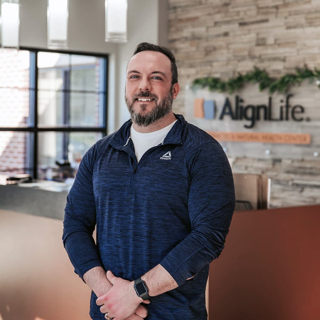 A man with a beard stands in a reception area near a counter, wearing a dark blue pullover and smartwatch. The background shows an "AlignLife" sign, highlighting the welcoming atmosphere of this Fishers Chiropractor office.