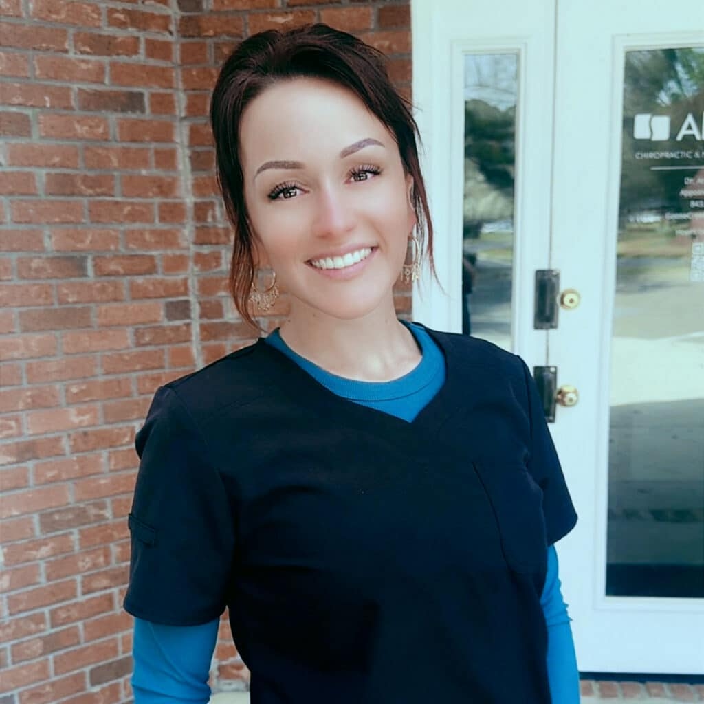 A woman wearing dark medical scrubs and a blue undershirt stands smiling in front of a brick building with glass doors, ready to welcome you as your trusted Goose Creek SC Chiropractor.