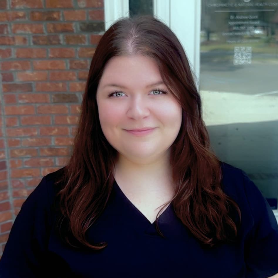 A woman with long brown hair wearing a dark scrub top stands outside in front of a brick wall and glass door, smiling at the camera—ready to welcome you as your friendly Goose Creek SC Chiropractor.