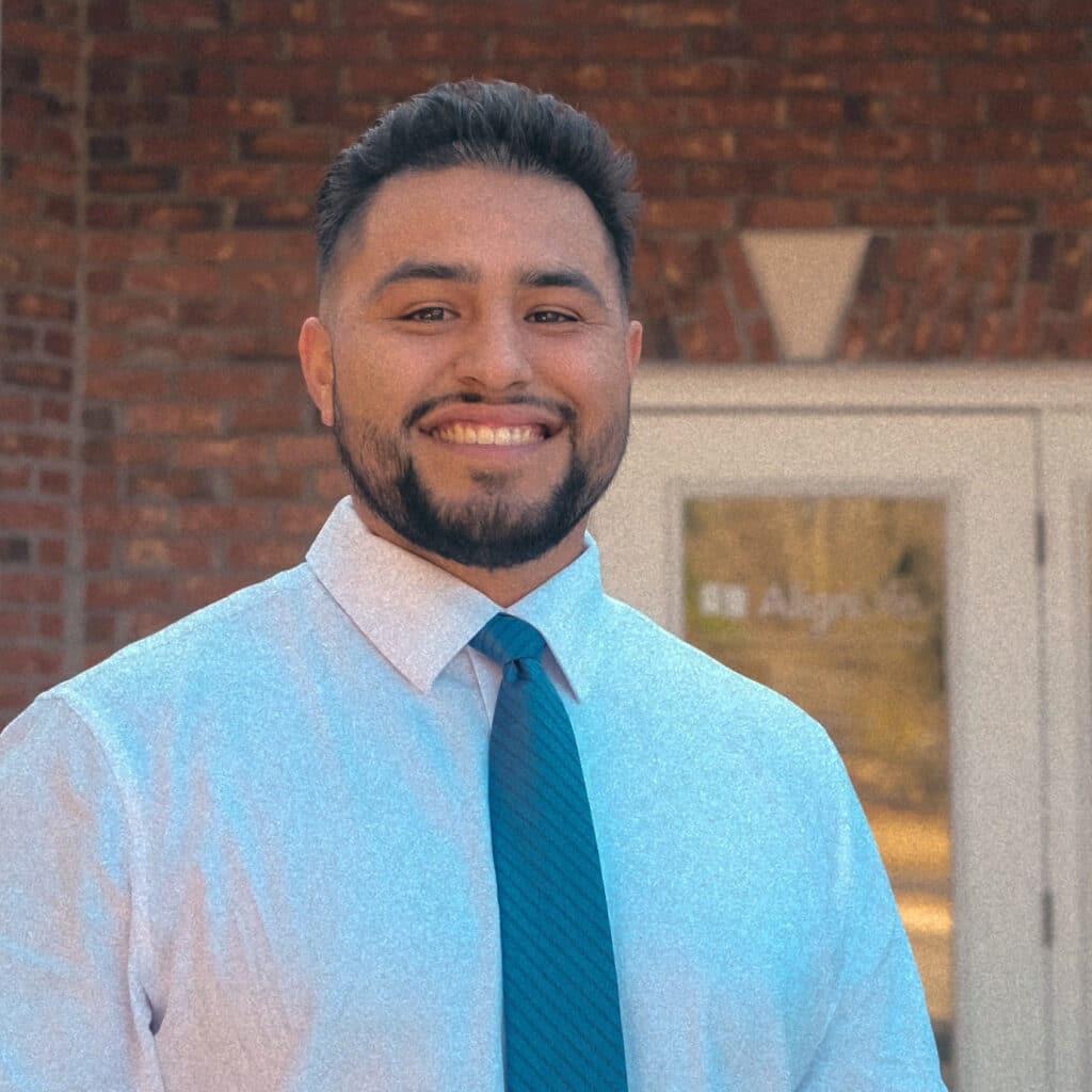 Dr. Joshua Martinez wearing a white shirt and teal tie, stands smiling in front of AlignLife Goose Creek, brick building with a glass door. Welcoming Goose Creek SC Chiropractor.