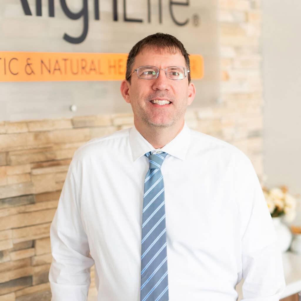 A man wearing glasses, a white shirt, and a blue striped tie stands in front of a stone wall with a partially visible Springfield Il Chiropractor business sign in the background.