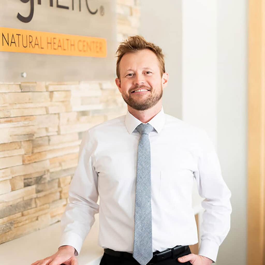 A man in a white dress shirt and tie stands in front of a wall sign that reads "NATURAL HEALTH CENTER," representing a trusted Springfield IL Chiropractor dedicated to wellness.