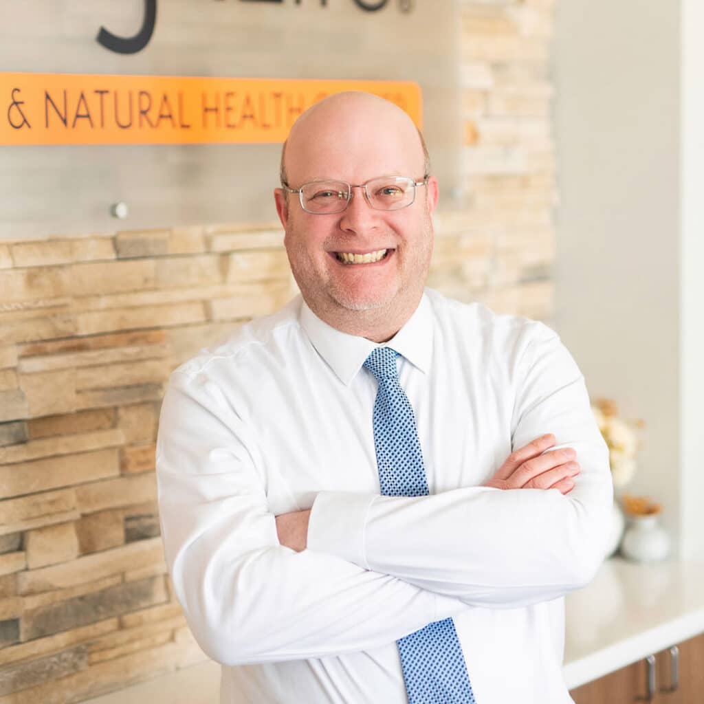 Bald man in glasses, a white shirt, and blue tie stands smiling with arms crossed in front of a stone wall and sign in an office setting, representing a friendly Springfield IL Chiropractor.