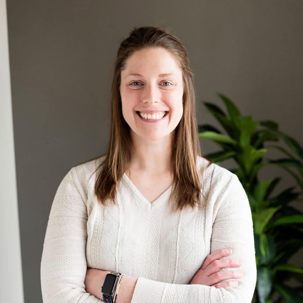 Woman with long brown hair in a white sweater stands with arms crossed, smiling, in front of a gray wall and green plant—capturing the welcoming vibe of a Springfield IL chiropractor’s office.