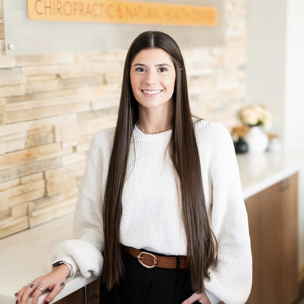 A woman with long brown hair, wearing a white sweater and black pants, stands indoors in front of a stone wall with a sign for a Springfield IL Chiropractor and natural health center.