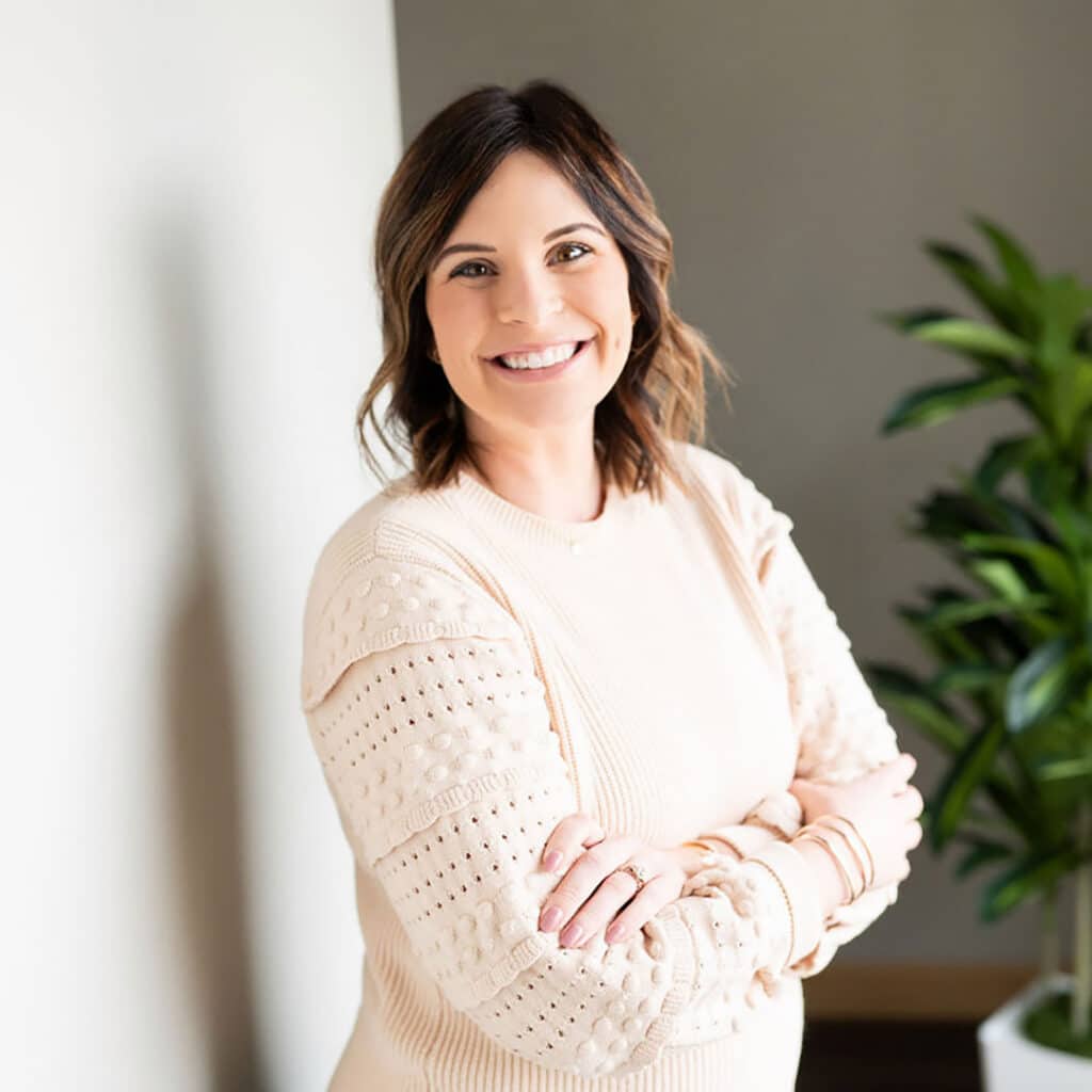 Woman with shoulder-length brown hair and a light textured sweater stands indoors with arms crossed, smiling at the camera; a green plant is visible in the background, reflecting the welcoming atmosphere of a Springfield IL chiropractor’s office.
