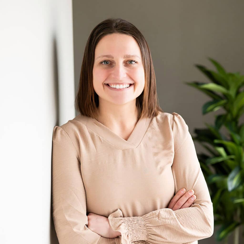 A woman with straight brown hair, wearing a beige long-sleeve blouse, stands with arms crossed and smiles at the camera—an inviting image for a Springfield IL Chiropractor. A green plant is visible in the background.
