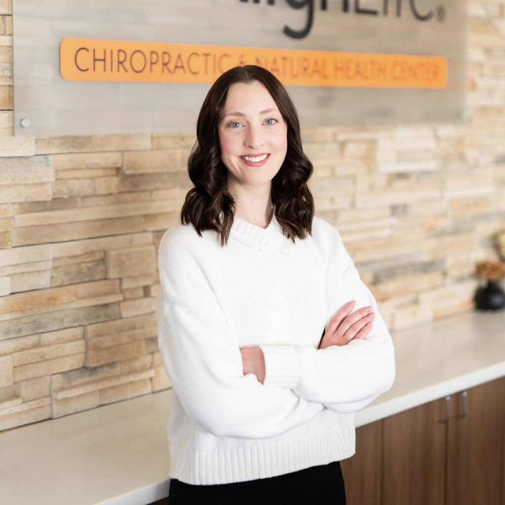 A woman with brown hair in a white sweater stands with arms crossed in front of a sign for a Springfield IL Chiropractor and natural health center.