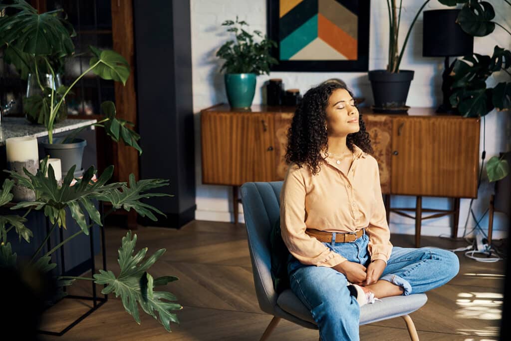 Woman sitting in living room chair enjoying the sunshine to relax, mediate and recharge from overall burnout - natural vitamin D