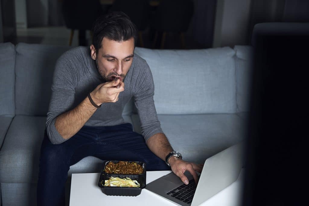 young man sitting on couch at night eating takeout food while working - factors that affect gut health