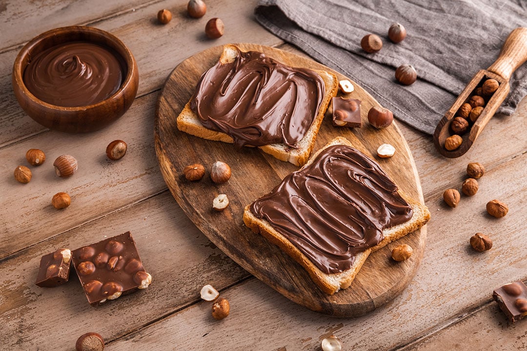 Board of bread with chocolate paste and hazelnuts on wooden background - Is Nutella Healthy