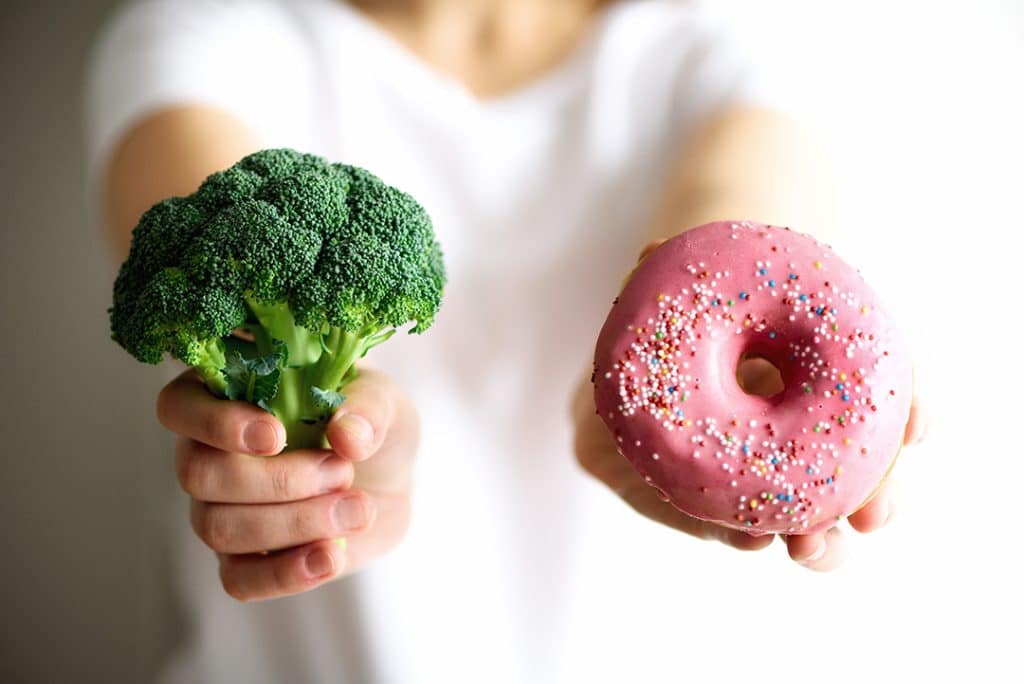 Young woman in white T-shirt choosing between broccoli or junk food, donut - Spring cleaning bad habits - developing healthy habits