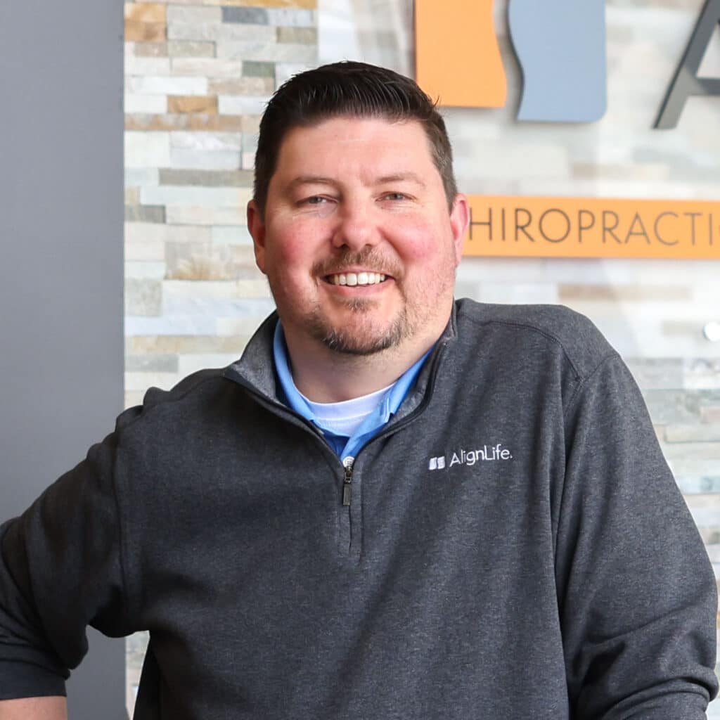 A man with short hair and a beard wearing an AlignLife pullover smiles while standing indoors in front of a stone wall and a Woodridge IL Chiropractor office sign.