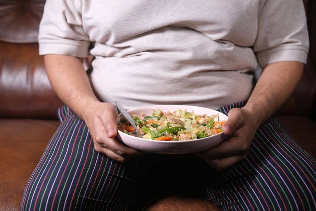 Overweight man sitting on couch eating a fresh healthy salad - inflammation, insulin resistance weight gain cycle