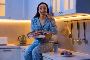 Young woman in pajamas eating pizza in kitchen at night. Eating habits for better sleep