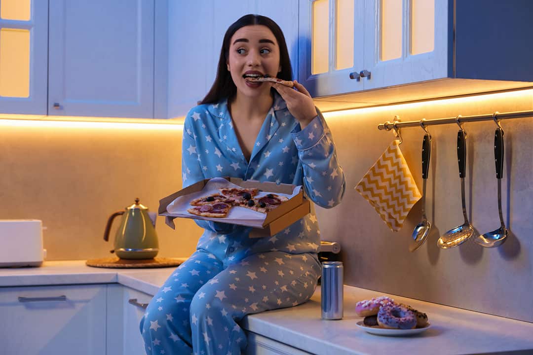 Young woman in pajamas eating pizza in kitchen at night. Eating habits for better sleep