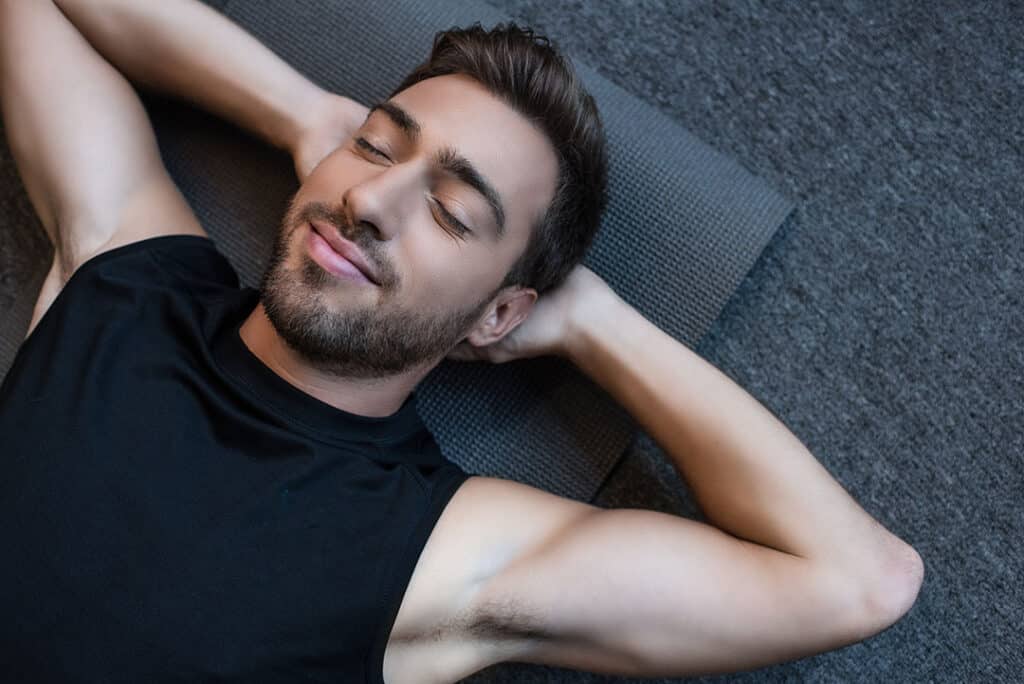 Young man in tanktop lying on a yoga mat with eyes closed and hands behind head - sleep vs exercise for weight loss