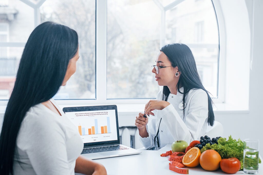 Female nutritionist with laptop gives consultation to patient indoors in the office.