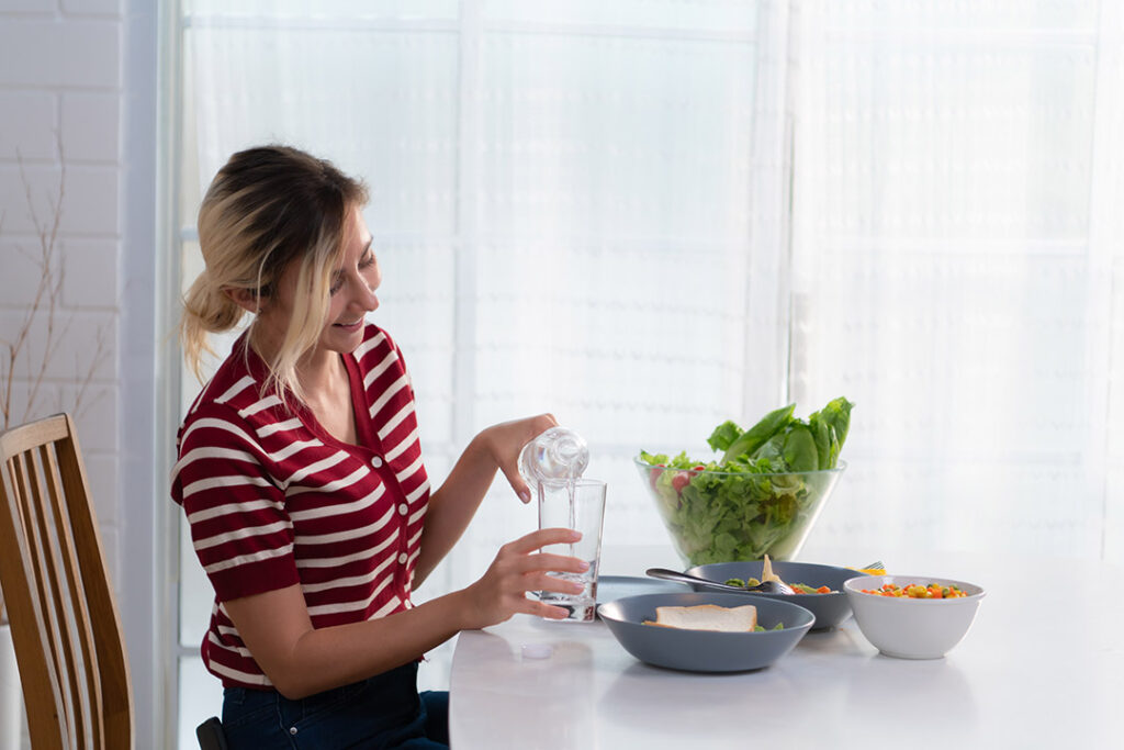 Woman pouring water into glass while sitting at a table - how to increase fiber in your diet