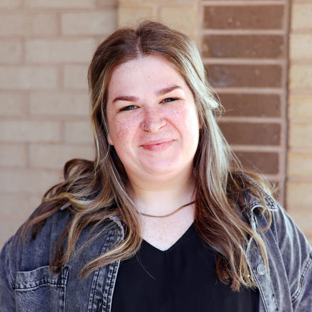 A woman stands front of a brick wall, smiling at the camera—capturing the friendly vibe of a Sugar Hill GA Chiropractor’s welcoming office.