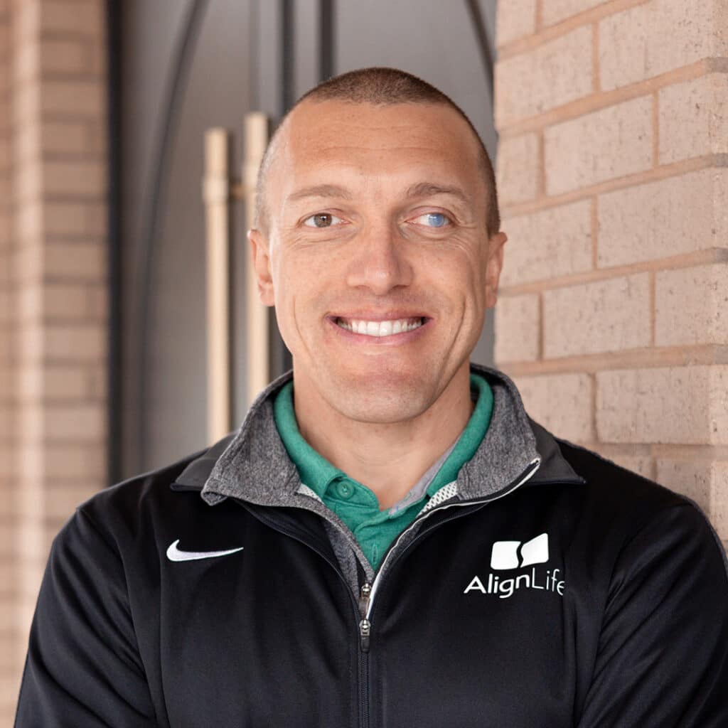 A doctor in an AlignLife jacket stands in front of a brick wall, smiling with his arms crossed—representing your trusted Sugar Hill Ga Chiropractor Dr. Dusti "Biggs" Biggerstaff