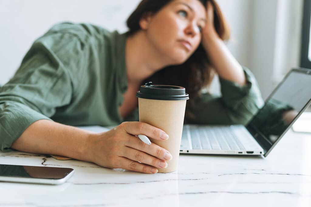 Young unhappy brunette woman plus size working at laptop on table with paper cup of coffee in bright modern office