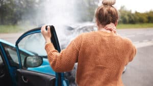 A woman stands outside a blue car with its door open, holding her neck after an apparent accident, suggesting possible personal injury, while smoke rises from the front of the vehicle.