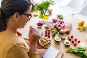 A woman sits at a table with a bowl of nuts, surrounded by various foods, a notepad, and pen, appearing to plan or record her meal choices during a healing crisis.