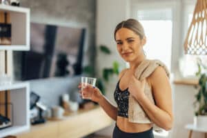 Woman in athletic wear holding a glass of water and a towel over her shoulder, standing indoors and looking at the camera—embodying the benefits of functional nutrition for an active lifestyle.