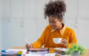 Woman in an orange sweater sits at a desk, holding a cup in one hand and writing on a tablet with a stylus, as she reviews notes on bone loss amid scattered papers and office supplies.
