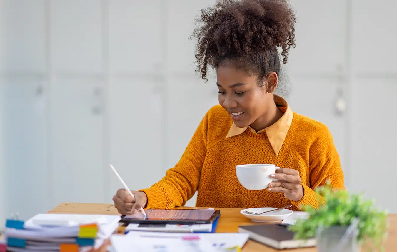 Woman in an orange sweater sits at a desk, holding a white mug and writing on a tablet with a stylus, surrounded by papers, a notebook, and a small plant—researching ways to prevent bone loss.