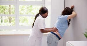 A healthcare professional examines a person standing and leaning against a wall, placing hands on the lower back to assess discomfort related to sciatica during pregnancy in a medical setting.