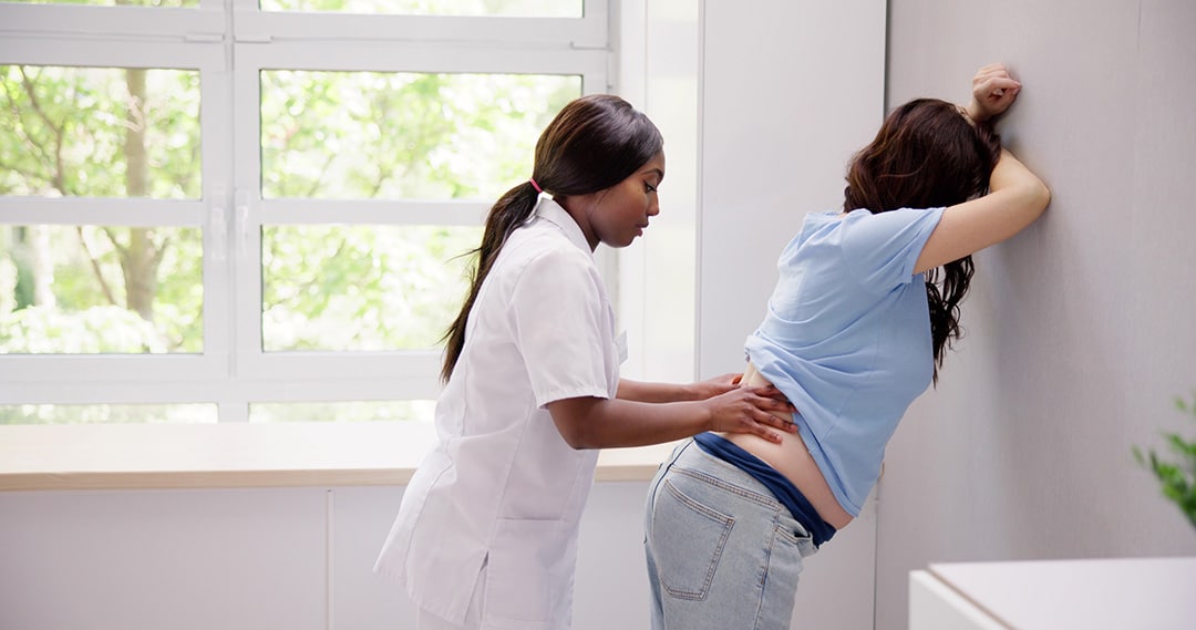 A healthcare professional examines a person standing and leaning against a wall, placing hands on the lower back to assess discomfort related to sciatica during pregnancy in a medical setting.
