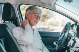 An older woman sitting in the driver’s seat of a car holds her neck, appearing to be in discomfort, possibly from whiplash.