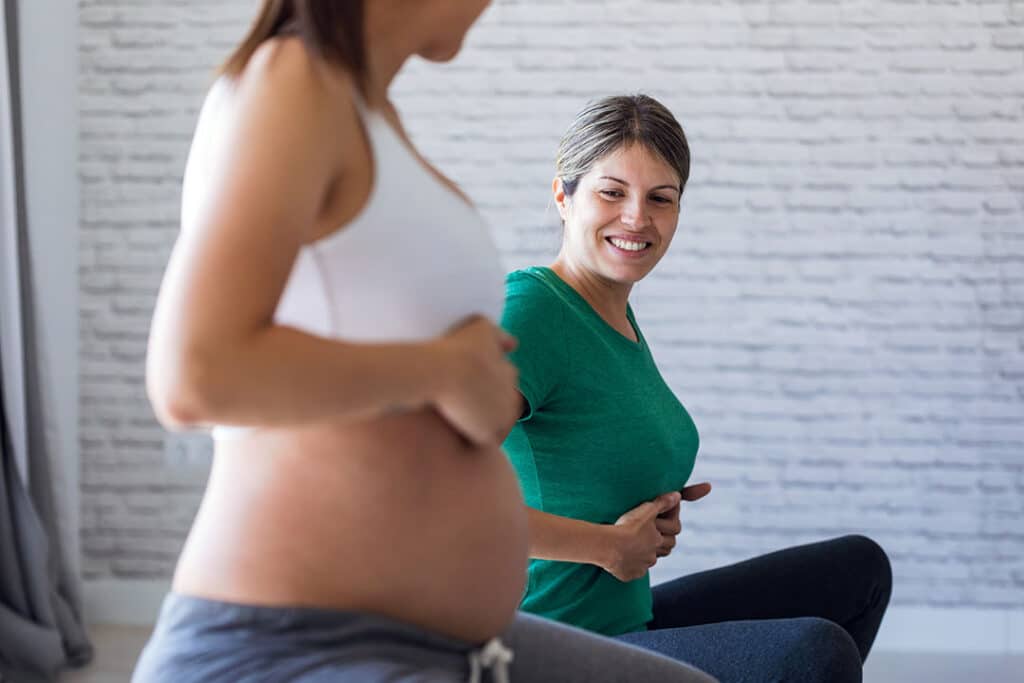 Two women are sitting indoors doing prenatal exercises; one, experiencing sciatica during pregnancy, is visibly pregnant while the other, in a green shirt, sits beside her and smiles supportively.