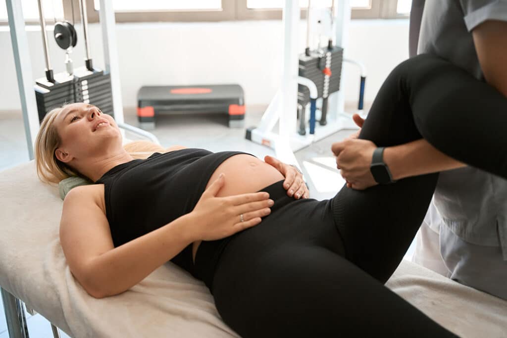 A pregnant woman lies on an exam table while a healthcare professional assists her in a leg exercise to help manage sciatica during pregnancy in a medical or physical therapy setting.