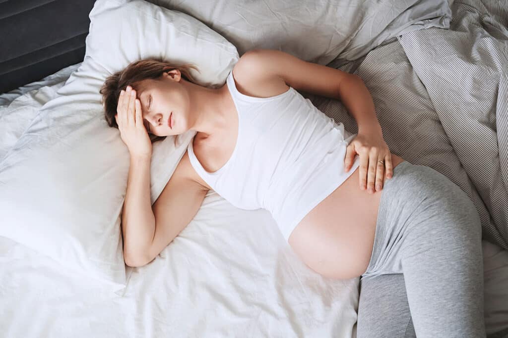 A pregnant woman lies on a bed, wearing a white tank top and gray pants, with one hand on her belly and the other on her forehead, appearing uncomfortable—possibly experiencing sciatica during pregnancy.