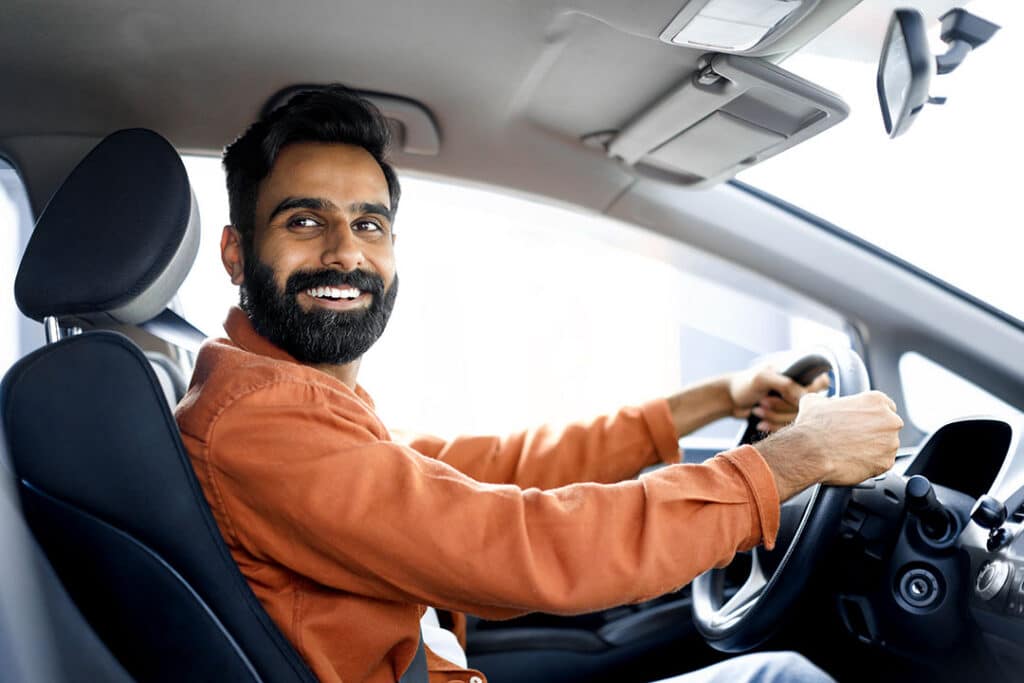 A man with a beard, wearing an orange shirt, sits in the driver's seat of a car, smiling and looking toward the camera—happy to be safe from whiplash on his journey.