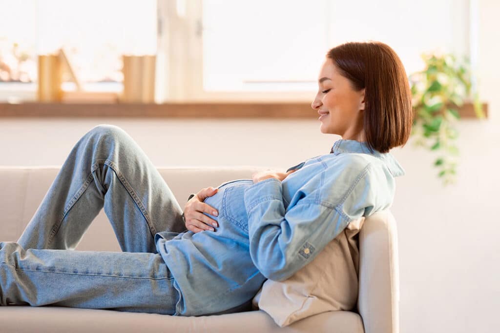 A pregnant woman in a denim shirt and jeans reclines on a couch, smiling and resting her hands on her belly in a bright, sunlit room—finding comfort even with sciatica during pregnancy.