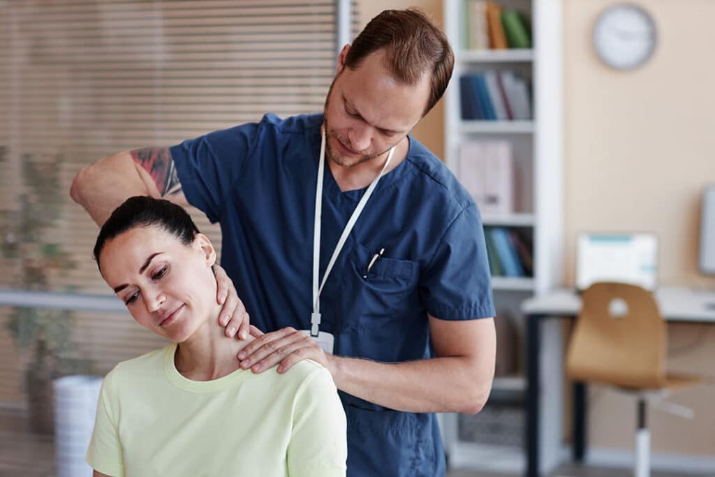 A healthcare professional in scrubs examines a woman’s neck for possible whiplash in a medical office. The woman is seated and tilting her head to the side during the assessment.