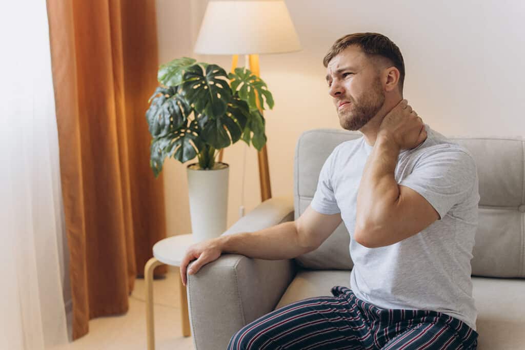 A man sitting on a couch holds his neck and looks uncomfortable, possibly experiencing neck pain from whiplash.
