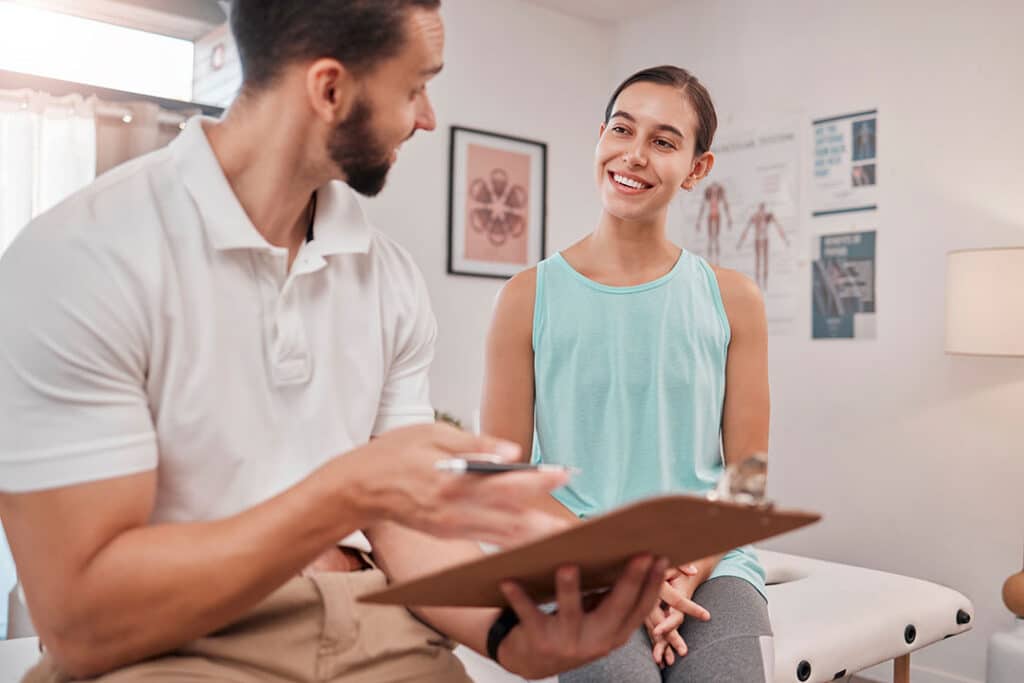 A man holding a clipboard talks to a smiling woman about whiplash treatment in a bright medical or therapy office setting.