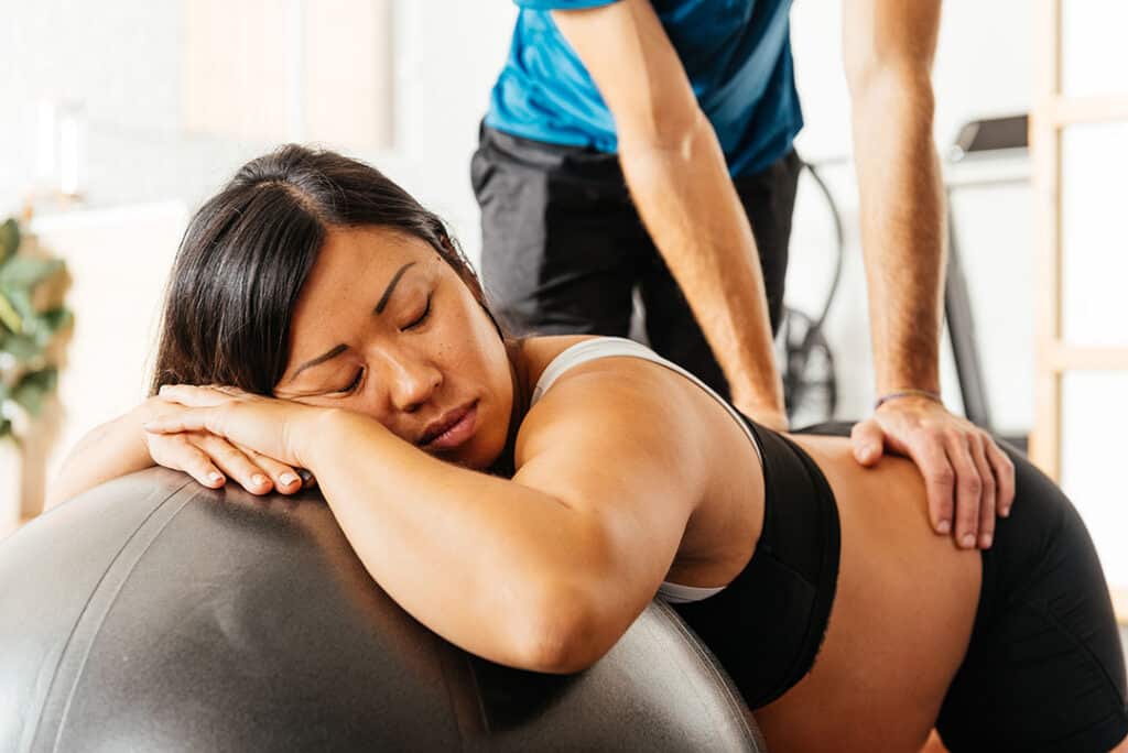 A woman experiencing sciatica during pregnancy rests her head on an exercise ball while a person massages her lower back.