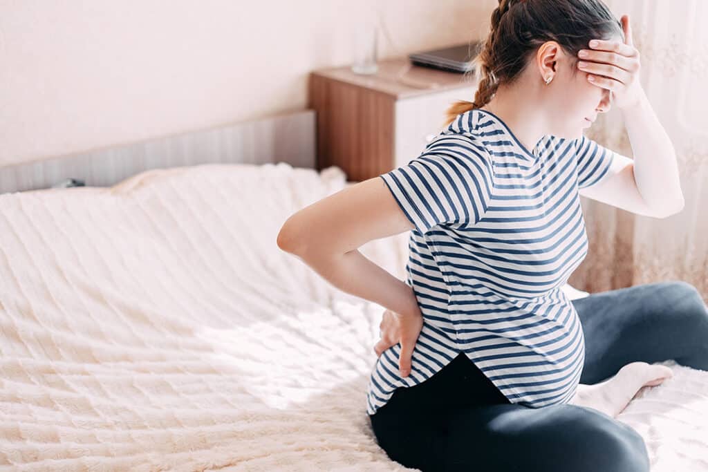 Pregnant woman sitting on a bed, holding her lower back with one hand and her forehead with the other, appearing to be in discomfort from sciatica during pregnancy.